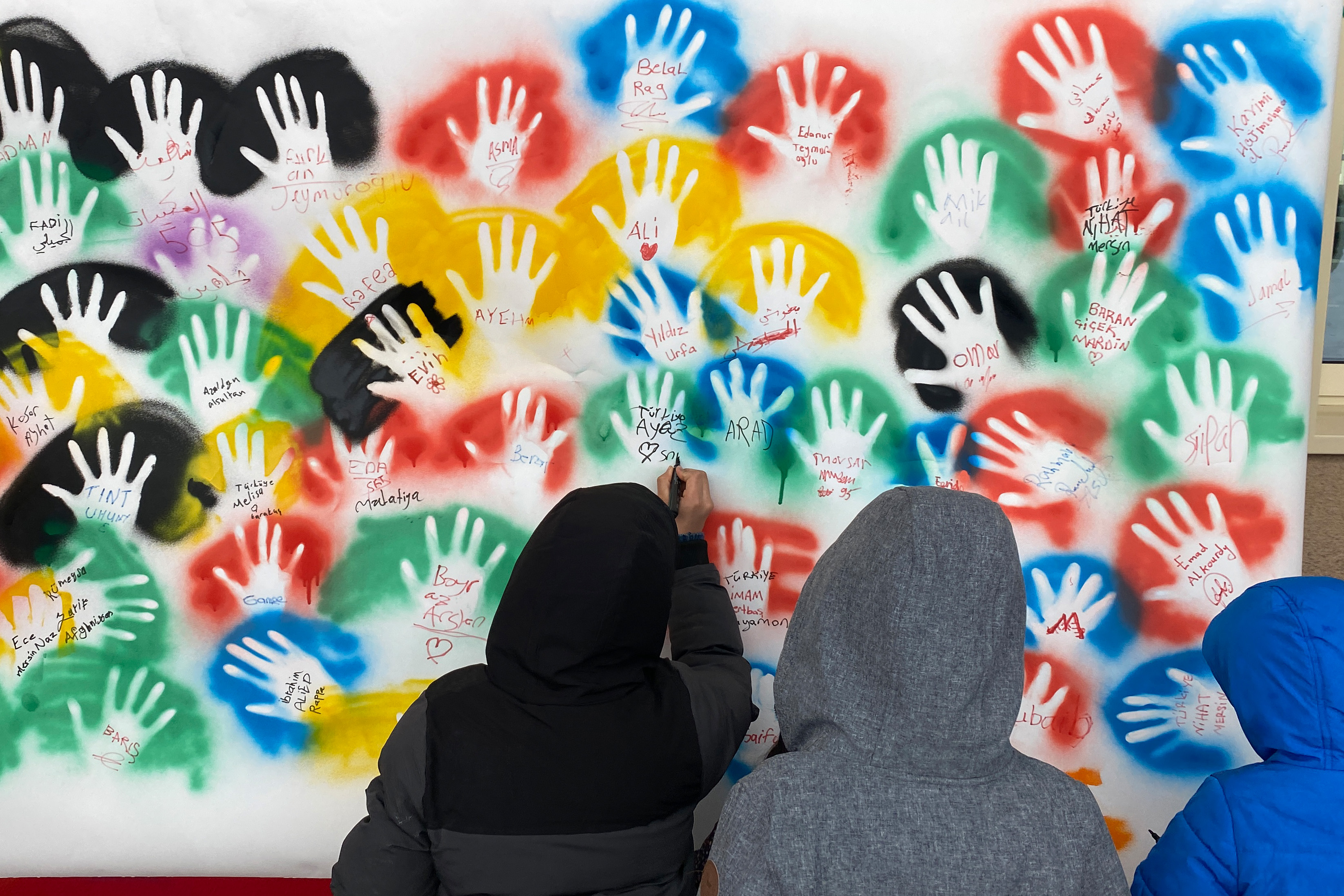 Drei Kinder stehen mit dem Rücken zur Kamera vor einer bunten Graffitiwand mit aufgesprühten Handabdrückenn